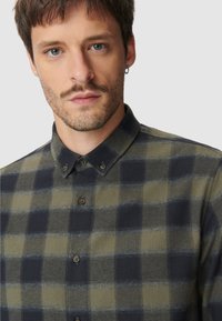 Jonge man met kort bruin haar en lichte stoppels, gekleed in een olijfgroen en zwart geblokt hemd met knopen, met een ringoorbel in één oor.