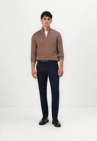 Hnedý sveter so zipsom v mäkkej tkanine, skombinovaný s tmavomodrými elegantnými nohavicami a čiernymi šnurovacími čižmami. Priehľadné okuliare dodávajú moderný vzhľad.