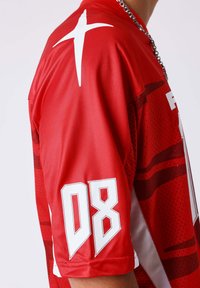 Personne portant un maillot de sport rouge avec le numéro blanc 08 sur la manche et un graphisme blanc ressemblant à une croix sur l'épaule, chaîne argentée visible.