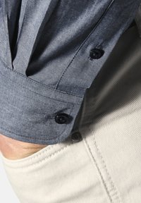 Kurzärmeliges Hemd aus dunkelblauem Stoff mit strukturierter Oberfläche, ausgestattet mit einem gefalteten Bündchen und zwei schwarzen Knöpfen, kombiniert mit hellen Hosen.