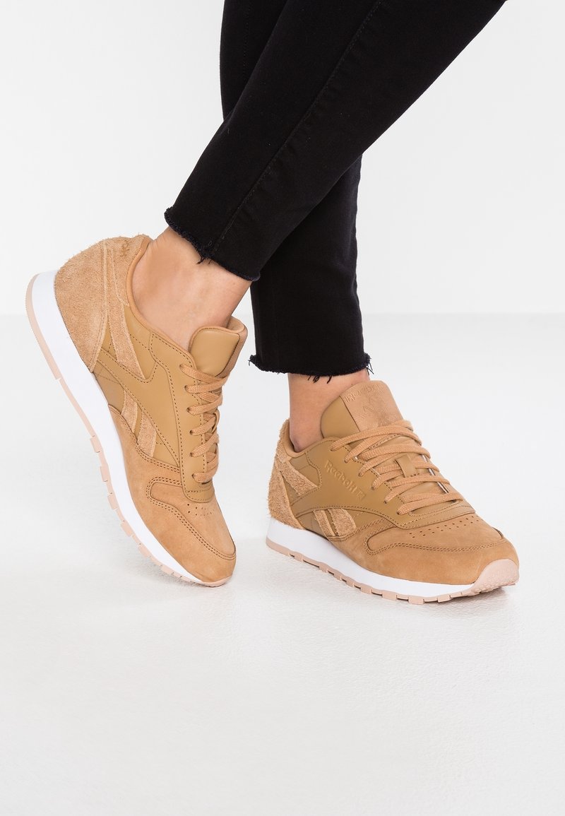 Person med beige mocka- och lädersneakers med vita sulor och svarta avkortade byxor, stående på ett vitt golv.