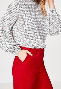 Biała bluzka w wzory z czarnym napisem i czerwonymi sercami, z bufkami na długich rękawach. Połączona z dopasowanymi czerwonymi spodniami, całość jest elegancka.