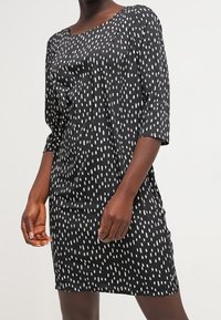 Robe noire à pois blancs verticaux, avec un décolleté bateau et des manches trois-quarts, fabriquée dans un tissu lisse, coupe décontractée.