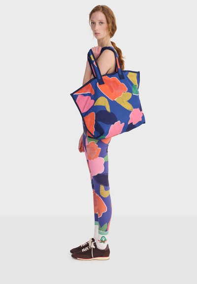 Jeune personne debout, portant des leggings floraux colorés et tenant un grand sac fourre-tout assorti à fleurs, porté sur une épaule, sur un fond uni.