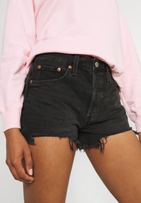 Shorts de mezclilla negra con dobladillo deshilachado, detalles desgastados, herrajes en tono cobre y diseño de cinco bolsillos. Combinados con un sudadera rosa clara.