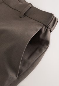 Gros plan de la ceinture d’un pantalon en tissu marron avec une boucle de ceinture et une poche latérale sur fond clair.