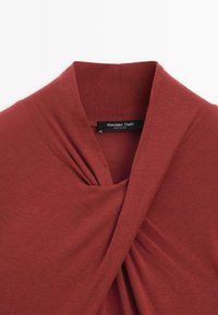 Červený pletený top s drapovaným výstřihem do V a detailním zasunutím v přední části. Tkanina je měkká, hladká a dokonale sedí, s viditelnou etiketou.