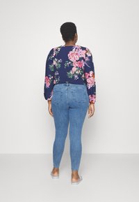 Blusa de manga longa com padrão floral sobre um fundo azul marinho, combinada com jeans claros ajustados, destacando o design nas costas e o corte slim e afunilado.