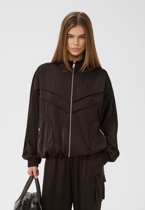 Jeune femme portant une veste noire à fermeture éclair et un pantalon assorti, tenant un sac à main texturé foncé sur un fond clair uni.