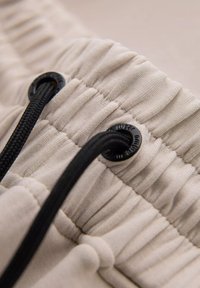 Bežová textilie se staženou strukturou; obsahuje černou šňůrku a plastové zámky na šňůrku pro nastavitelnou velikost.