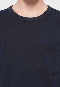 Navy sininen puuvillapaita, jossa on pyöreä kaula-aukko, lyhyet hihat ja rintatasku. Kankaan pinta on sileä ja siinä on hienoinen rakenne.