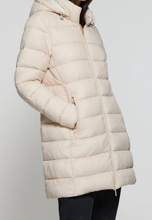 Kobieta ubrana w długi, beżowy, pikowany płaszcz puchowy z kapturem i kieszeniami na zamek, stojąca z jedną ręką w kieszeni.