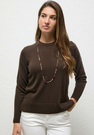 Joven mujer con cabello largo y castaño lleva un suéter marrón oscuro, pantalones blancos y un collar largo con cuentas rojas y doradas sobre un fondo liso.