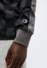 Χέρι με σκουρόχρωμο δέρμα που φοράει μακρυμάνικο φούτερ σε μαύρο και γκρι, με μικρό κεντημένο λογότυπο Champion σε μπλε και κόκκινο κοντά στον καρπό.