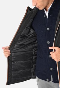 Giubbotto piumino nero con cerniera arancione, caratterizzato da un interno trapuntato e un maglione lavorato a maglia blu navy con dettagli sui bottoni sotto.
