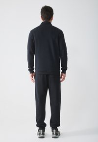 Set de hanorac și pantaloni sport negri, realizat dintr-un material moale. Prezintă manșete ribbate și o croială lejeră. Vedere din spate arată detalii minime.
