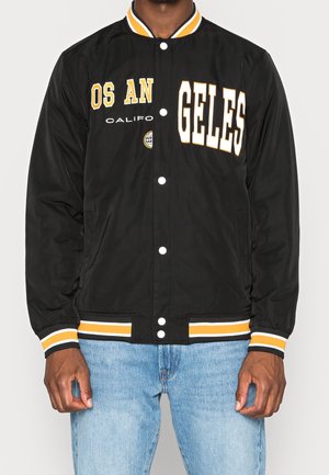 Homme portant une veste varsity noire avec des bordures jaunes et blanches, texte "LOS ANGELES CALIFO" à l'avant, associée à un jean bleu clair.