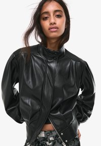 Černá kožená bunda s vysokým límcem, předními patentními knoflíky a zipem na spodním okraji. Texturovaný povrch s nařasením na ramenou.