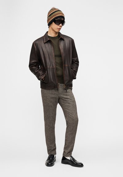 Άνδρας που φοράει καφέ δερμάτινο μπουφάν, πράσινο πουλόβερ, γκρι υφασμάτινο παντελόνι, πλεκτό καπέλο, μαύρα γυαλιά ηλίου και μαύρα μοκασίνια, στέκεται με τα χέρια στις τσέπες.