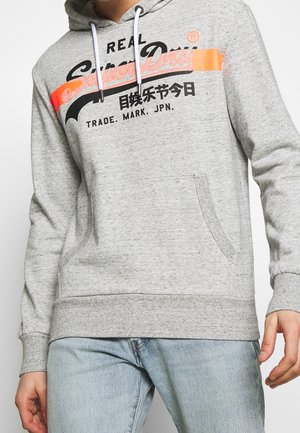 Osoba ubrana w jasnoszarą bluzę z kapturem SuperDry z pomarańczowo-czarnym logo oraz jasnoniebieskie dżinsy, stojąca na białym tle.