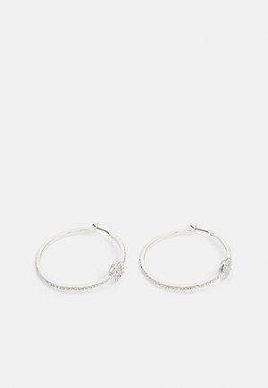 Paire de boucles d'oreilles créoles argentées avec petits cristaux incrustés et un plus grand détail en cristal rond près du fermoir sur fond blanc.