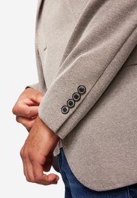 Helles beige Blazer aus strukturiertem Stoff, mit vier schwarzen Knöpfen an den Ärmeln und taillierter Passform, getragen über einem weißen Hemd und blauen Jeans.