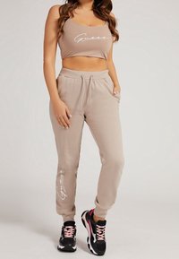 Beżowy krótki top z białym logo "Guess" w zestawie z dopasowanymi spodniami dresowymi z elastycznym pasem, bocznymi kieszeniami i detalem z logo.