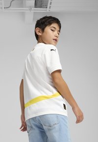 Balts īsām piedurknēm sporta krekls ar dzeltenu joslu jostā, ar Puma logotipu uz pleca. Valkāts ar gaiši zilo džinsu biksēm.