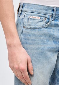 Ανοιχτό μπλε τζιν Calvin Klein με ορατή ετικέτα μάρκας στην μικρή μπροστινή τσέπη, φορεμένα από ένα άτομο με λευκό τοπ και το χέρι του χαλαρό δίπλα στο σώμα.