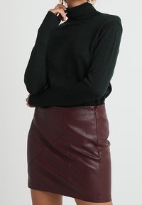 Dunkelgrüner Rollkragenpullover mit langen Ärmeln, kombiniert mit einem burgunderfarbenen Leder-Minirock, der diagonale Nahtdetails und einen Reißverschluss hat.