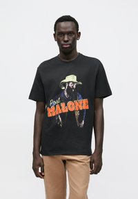 Tricou negru din bumbac, cu o imprimare grafică colorată a unui bărbat cu pălărie de cowboy, cu textul "Post Malone" în portocaliu. Mâneci scurte, guler rotund.