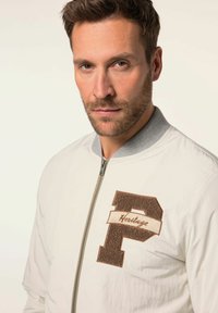 Giacca bomber bianca con colletto grigio a costine, caratterizzata da una toppa di feltro marrone con la lettera "P" e la scritta "Heritage". Chiusura con zip, texture liscia e design aderente.
