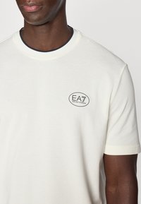 Μπεζ μπλουζάκι με κοντά μανίκια, ριμπ navy γιακά και μικρό οβάλ λογότυπο σε σκούρο μπλε, με κείμενο "EA7 Emporio Armani".