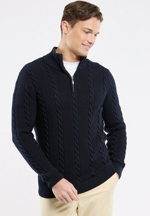 Homme portant un pull à torsades bleu marine avec col demi-fermeture éclair et pantalon beige, souriant et regardant vers le bas, les mains dans les poches.