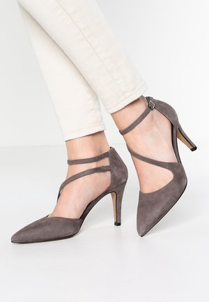 Vrouw die grijze suède pumps met spitse neus en enkelbandjes draagt, gecombineerd met off-white broek tegen een effen achtergrond.