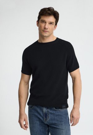 Mand står iført en sort kortærmet striktrøje og blå jeans mod en ensartet lys baggrund.