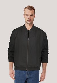 Schwarze Bomberjacke mit geripptem Kragen und Bündchen, Reißverschluss vorne und glatter Textur. Über blauen Jeans getragen.