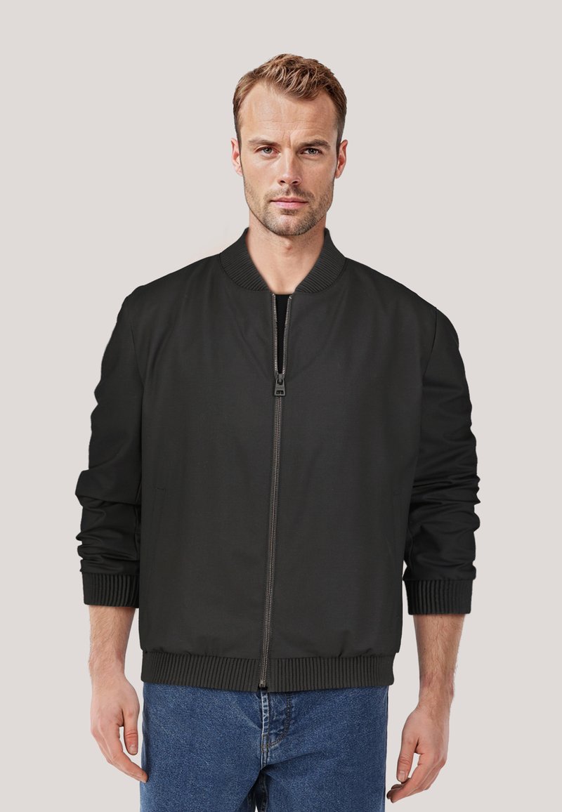 Schwarze Bomberjacke mit geripptem Kragen und Bündchen, Reißverschluss vorne und glatter Textur. Über blauen Jeans getragen.