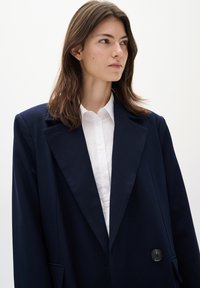 Blazer oversize blu navy con rever affilati, chiusura a un bottone e tasche; abbinato a una camicia bianca con colletto sotto.