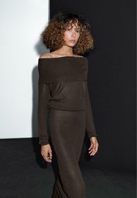 Frau mit lockigem Haar, die ein braunes, schulterfreies, geripptes Stricktop und einen passenden langen Rock vor einem Schwarz-Weiß-Hintergrund trägt.