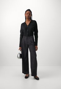 Sort tekstureret skjorte med knapper, grå højtaljede plisserede bukser, sorte flade sko, holder en sølvfarvet håndtaske, minimalistisk baggrund.