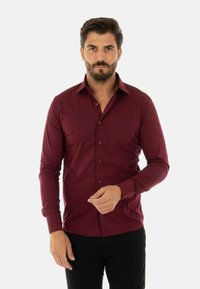 Camicia a maniche lunghe bordeaux con un design aderente, colletto button-down e texture liscia. Abbinata a pantaloni neri, con una leggera lucentezza.