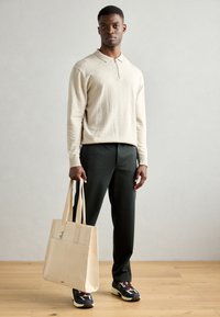 Pull en beige à col zippé, pantalon vert foncé et sac tote en toile. Baskets avec accents blancs et navy. Textures lisses, couleurs contrastantes.