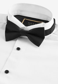 Camisa blanca de vestir con tejido texturizado, que presenta una pajarita negra, botones negros y un patrón de lunares en el interior del cuello.