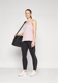 Lys rosa treningstopp, svarte leggings med hvite sidestreker, og hvite sneakers med lilla detaljer. Holder en svart treningsbag.