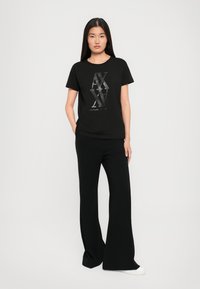 Naine seisab käed taskus, kandes musta Armani Exchange logo t-särki, musti laiad püksid ja valgeid spordijalgu valgel taustal.