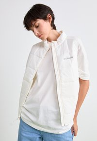 Jong persoon met kort, zwart haar die een witte Columbia gewatteerde vest over een wit T-shirt en lichte blauwe spijkerbroek draagt, tegen een effen achtergrond.