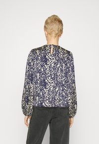 Persona con capelli biondi corti indossa una blusa con maniche lunghe di colore navy e beige a fantasia e jeans grigi scuri, mostrata di spalle su sfondo bianco.
