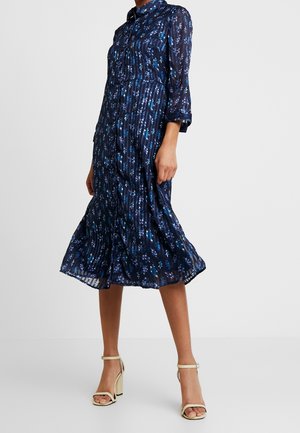 Mujer que lleva un vestido midi floral azul marino de manga larga con detalles de botones, combinado con sandalias blancas de tacón alto con tira en el tobillo.