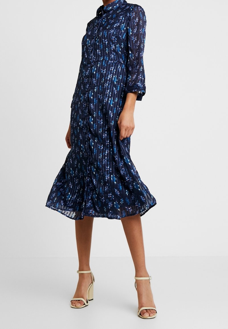 Femme portant une robe midi fleurie bleu marine à manches longues avec des boutons, associée à des sandales blanches à talons hauts avec bride à la cheville.